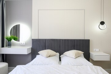 modern bedroom featuring a gray upholstered headboard, white linens, a round lighted mirror, and...