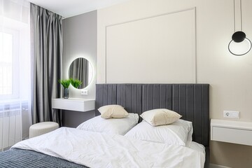 modern bedroom featuring a gray upholstered headboard, white linens, a round lighted mirror, and...