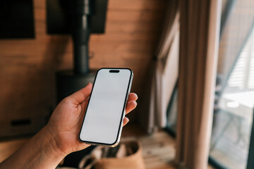 Person holding a smartphone with blank white screen in front of a modern wood stove, ready for app...