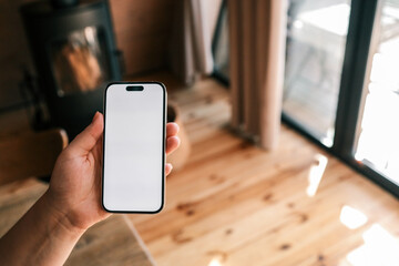 Person holding a smartphone with blank white screen in front of a modern wood stove, ready for app mockup or design presentation in a cozy interior