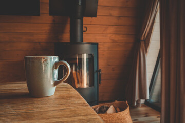 Ceramic mug on a wooden table in front of a modern black wood stove, cozy interior with natural light and warm wooden tones