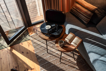 Top-down view of a cozy reading and music corner in a sunlit cabin, featuring an open book, cup of coffee, and a vinyl record player on wooden side tables near a gray sofa © shine.graphics