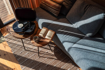 Top-down view of a cozy reading and music corner in a sunlit cabin, featuring an open book, cup of coffee, and a vinyl record player on wooden side tables near a gray sofa