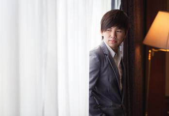 The younng asian businessman is standing nearby the window with white curtain.