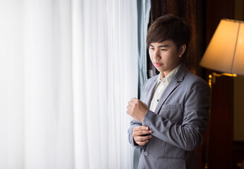 The younng asian businessman is standing nearby the window with white curtain.