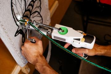 Close-up of a hand-held tufting gun being used to create a rug with green yarn, showing detail of...