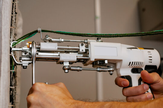 Close-up of a hand-held tufting gun being used to create a rug with green yarn, showing detail of industrial textile equipment and DIY carpet making process