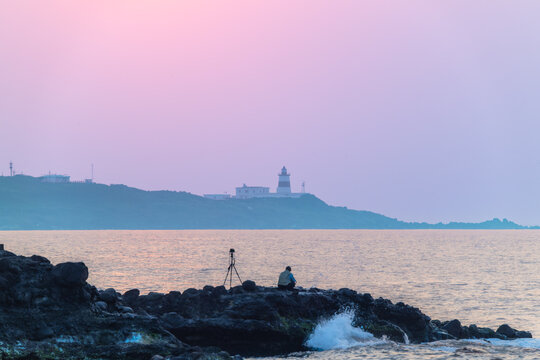 A lone photographer with a tripod captures the serene sunset over the Shimen coastline in New Taipei City, Taiwan, with the iconic Fuguijiao Lighthouse in the distance.