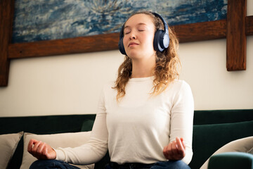 Young woman meditating with closed eyes and headphones on sofa in living room at home, practicing...