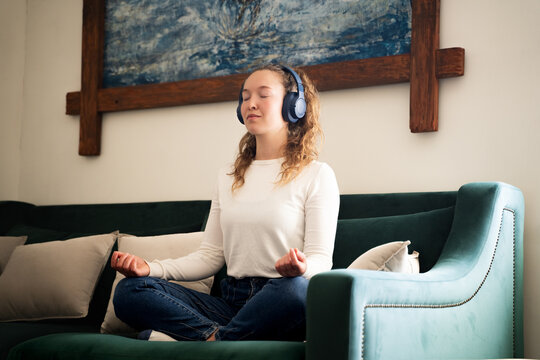 Young woman meditating with closed eyes and headphones on sofa in living room at home, practicing mindfulness, listening to music or guided meditation, mental health and relaxation, self care routine