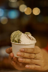 Hand holding a cup of ice cream with two scoops, one green and one white with blurred background
