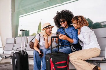 Multi ethnic friends sitting on airport bench with luggage, looking at smartphone and expressing...