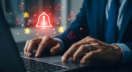 Man in suit working on laptop with red alert bells and warning signs floating above, indicating urgent notifications or cybersecurity issues.