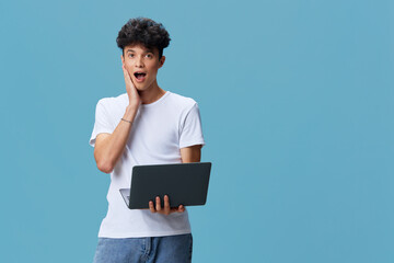 surprised young man holding laptop with open mouth and hand on cheek, wearing white t-shirt and...