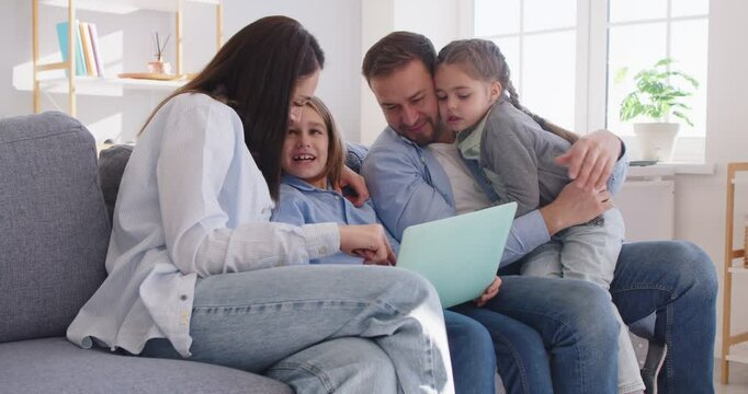 Happy family at home using laptop, cheerful parents, little kids, happy mom and dad enjoying leisure at home together, using computer on sofa, laughing, watching movie, interactive online TV channel