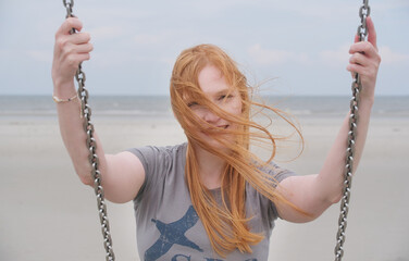 A red-haired woman on a swing at the beach, wind tousling her hair as she smiles with a sense of...
