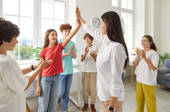 Joyful preteen school children celebrating successful teamwork result with their young friendly teacher. Female tutor and happy girl giving high five to each other with classmates applauding around.