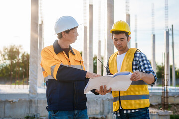 An elderly man and a middle-aged Asian man, both wearing hard hats and reflective vests, closely...