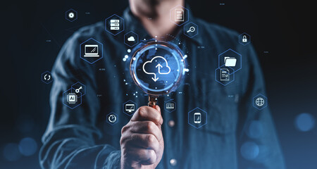 Man holding magnifying glass over cloud computing icon with futuristic digital interface and data technology symbols on dark blue background concept..