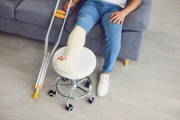 Handicapped man sitting on sofa at home on rehabilitation with crutches and with leg in plaster...