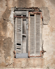 An old wooden window shutter with flaking white paint and rusting brackets on a crumbling old building at Girolata on the Mediterranean island of Corsica