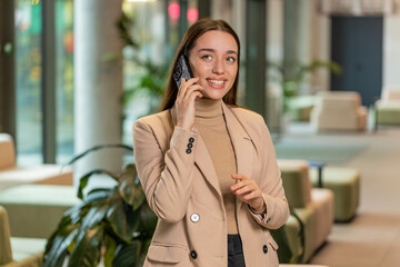 Smiling young businesswoman in formal working, having mobile phone talk in modern office lobby....