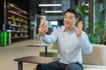 Portrait of smiling young businessman holding smartphone waving hand sign hello greeting...