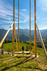 Fototapeta premium Girl on giant swing overlooking mountain valley. Young girl sits on a massive wooden swing, facing a stunning mountain landscape under a bright blue sky in summer.
