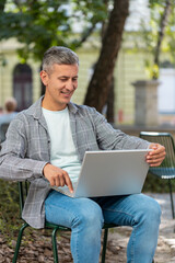 Happy mature man opens laptop sitting on chair in urban city street outdoors. Caucasian guy working...