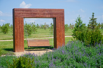A wooden bench swing hanging by metal chains from a large timber frame in a sunny urban park, surrounded by green grass, purple blossoms, and young trees. Photo