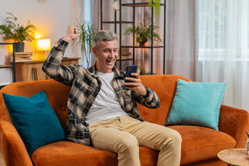 Surprised excited young man winner holding smartphone reading good news amazed by online bet bid game win. Happy guy male on sofa looking at screen overjoyed by victory success in home apartment.