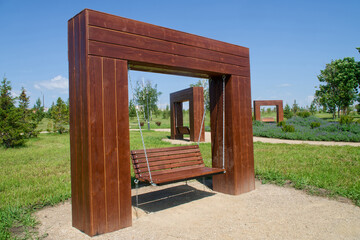 A large wooden swing bench suspended by metal chains from a sturdy timber frame in a green park, with additional swing installations receding into the background. Photo