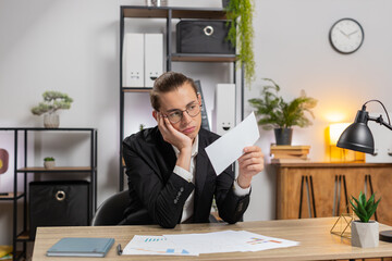Serious Caucasian young businessman in formal suit sitting at home office with letter, upset and...