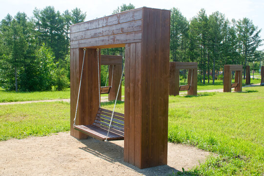 A series of wooden swing benches suspended by metal chains from large timber frames in a sunlit urban park, surrounded by grass and trees. Photo