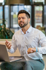 Sad nervous young businessman waiting to distance remote meeting conference with someone on laptop in modern office lobby. Upset guy looking at watch and looking at screen. Lack of punctuality concept