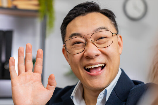 POV of excited Asian middle-aged businessman blogger influencer making phone conversation call with social media followers at office workplace working on laptop. Freelancer Chinese man in formal suit