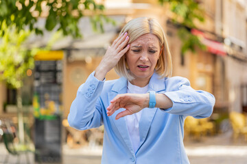 Shocked mature woman businesswoman in hurry checking time on smartwatch walking on city street. Medium shot of Caucasian middle-aged female entrepreneur late for business meeting walk fast outside.