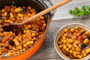 A cast-iron pot with chickpea and aubergine stew and a wooden spoon in it. A ceramic bowl of stew and parsley sprig. Rustic wooden table.