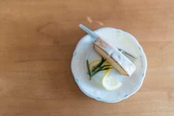 A slice of vanilla and lemon layer cake with white icing and lemon slice, served on a white plate with a fork beside it.
