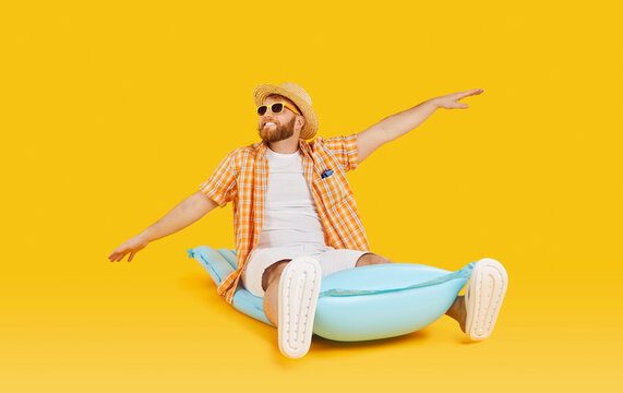 Happy smiling bearded young man wearing summer shirt, sunhat and sunglasses sitting on water mattress and holding arms outstretched on yellow orange color studio background. Holiday, vacation concept