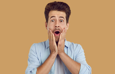 Portrait of young amazed scared man in blue shirt on beige background looking at camera. He...