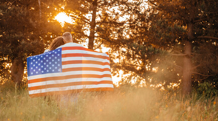A couple embraces peacefully in a sunlit meadow wrapped in an American flag, surrounded by tall grass and glowing trees in the warm golden light of a summer sunset