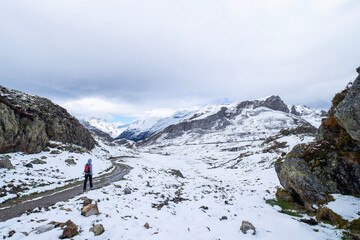unrecognizable alone hiker with a red backpack walks along a snow-covered mountain trail surrounded by rocky peaks under a cloudy sky in a serene winter landscape