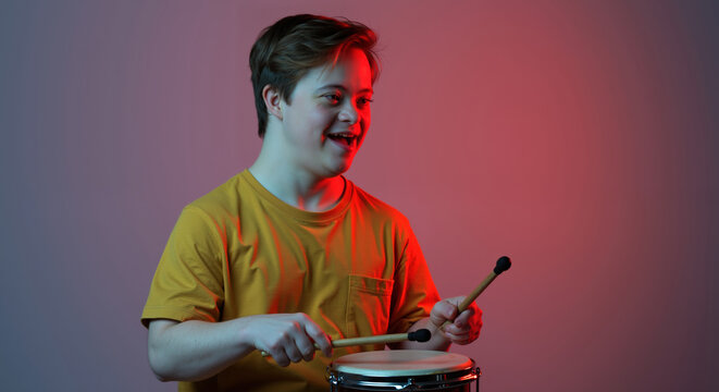 Smiling boy with down syndrome playing drums with sticks, yellow shirt, colorful gradient lighting. Special needs child musical performance, inclusive arts concept