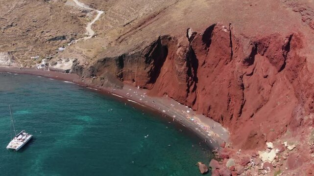 Aerial footage capturing the iconic Red Beach (Kokkini Paralia) in Santorini, Greece, showcasing its striking red volcanic cliffs, dark sand, and the vibrant turquoise Aegean Sea.