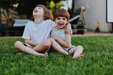 Two happy boys laughing together while sitting on the grass, enjoying a sunny day. Their playful...