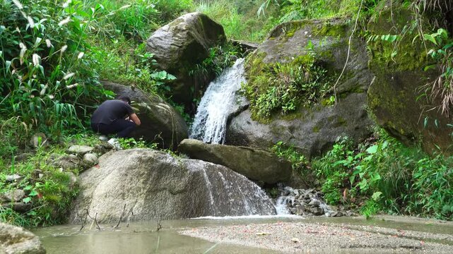 A man photographs a small stream and waterfall in a green forest setting. Ideal for editorial use depicting outdoor exploration and nature photography.