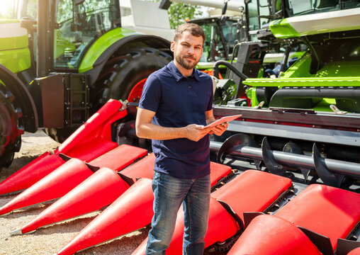 Man sales representative with digital tablet standing near combine harvester at agricultural machinery dealership.