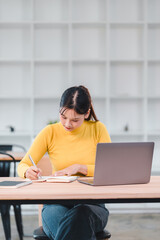 Young woman writing in notebook with laptop on wooden table in modern room, focused student studying indoors with casual outfit and natural light