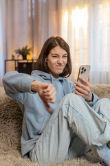 Dislike. Upset Caucasian woman using smartphone sitting on sofa couch at home showing thumbs down...